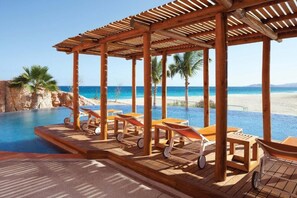 Ett trädäck vid poolen med solstolar och en pergola med utsikt över en strand och havet.