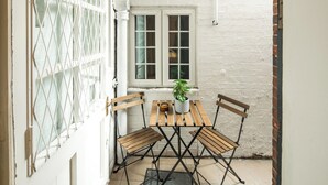 A small patio with a wooden table and chairs, a potted plant, and a window with white shutters. - Central London Gem | Sleeps 8 | Hyde Park | London (London)