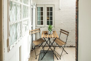 Un pequeño patio con mesa y sillas de madera, una planta en maceta y una ventana con contraventanas blancas.