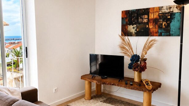 Ein modernes Wohnzimmer mit einer Couch, einem Fernseher auf einem Holzboden, einem Bild an der Wand und Blick durch eine Glastür.