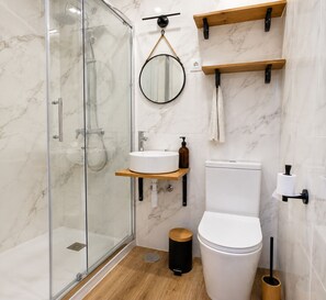 Et moderne badeværelse med brusekabine i glas, et hvidt toilet, et rundt spejl og træhylder.
