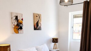 Ein ordentlich bezogenes Bett mit weißen Leinen, eine Nachttischlampe und zwei abstrakte Gemälde an der Wand.