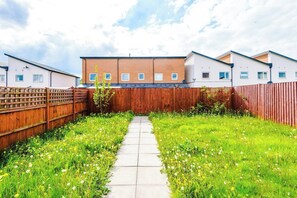 A residential backyard with a paved pathway, wooden fence, and a variety of flowering plants.