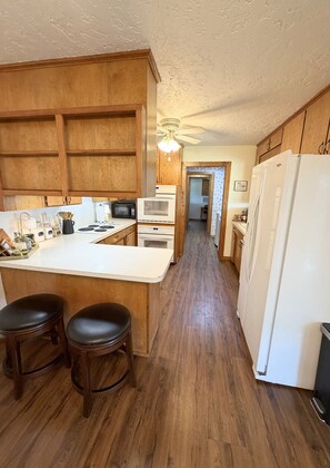 A kitchen with wooden cabinets, a white countertop, two stools, a refrigerator, and a microwave.