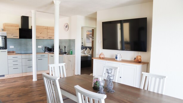 A modern kitchen with white cabinets, a wooden dining table, and a flat-screen TV mounted on the wall. - Ullrichshof Vacation Rental in Upper Lusatia – Sauna, BBQ Area & Panoramic View (Großdubrau)