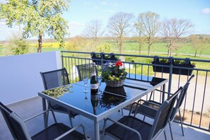 A balcony with a glass table, chairs, a bottle of wine, and a basket of flowers. - Ullrichshof Vacation Rental in Upper Lusatia – Sauna, BBQ Area & Panoramic View (Großdubrau)