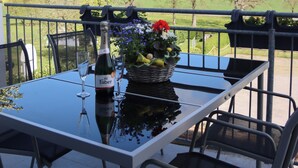 A table with a bottle of wine, glasses, and a basket of flowers on a balcony overlooking a green landscape. - Ullrichshof Vacation Rental in Upper Lusatia – Sauna, BBQ Area & Panoramic View (Großdubrau)