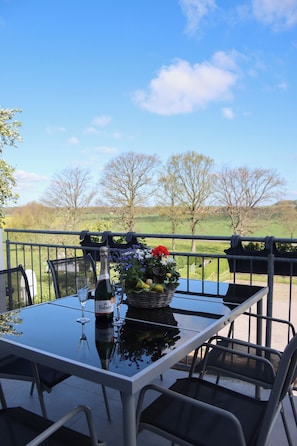 Een tafel met een fles wijn, glazen en een mand met bloemen op een balkon met uitzicht op een groen landschap.