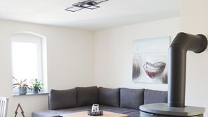A modern living room with a grey sofa, a white coffee table, and a black wood-burning stove. - Ullrichshof Vacation Rental in Upper Lusatia – Sauna, BBQ Area & Panoramic View (Großdubrau)