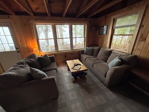 A cozy cabin interior with a wooden ceiling, a large window with a view of trees, a brown sofa, and a small wooden table.
