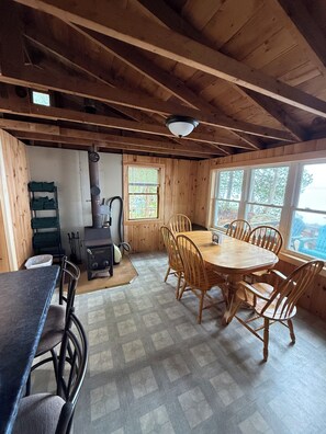 A wooden cabin interior with a dining table, chairs, a stove, and a window overlooking a forest.