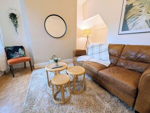 Ein Wohnzimmer mit braunem Ledersofa, einem runden Spiegel, einem kleinen Tisch mit einer Pflanze und einem Sessel.