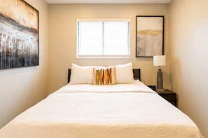 Habitación de hotel con cama, ventana con persianas, mesita de noche con lámpara y cuadros en las paredes.