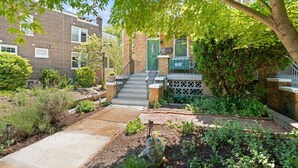 Un immeuble résidentiel à la façade en briques, avec une porte verte, un escalier menant à un porche et un jardin agrémenté de diverses plantes et d’un élément décoratif en roche.