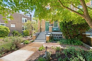 A residential building with a brick facade, a green door, a staircase leading to a porch, and a garden with various plants and a rock feature. - Hill East, Barney Circle 2-BR, Walk to METRO Stop (Washington)