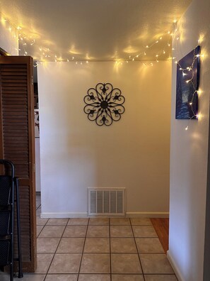 Una habitación con un adorno en la pared, luces de cuerda, una rejilla de ventilación y una silla.