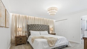 Ein Schlafzimmer mit einer gesteppten Kopfteil, einem Bett mit weißer Bettwäsche, zwei Nachttischlampen, einem Nachttisch aus Holz und einem großen Fenster mit durchsichtigen Vorhängen.