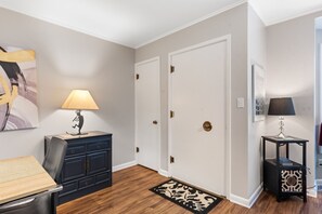 Un couloir avec un bureau, deux portes, une lampe, un tapis et un tableau accroché au mur.
