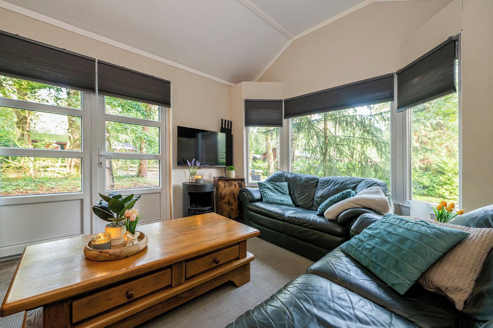 Een moderne woonkamer met een houten salontafel, een zwarte leren bank en grote ramen met uitzicht op groen.