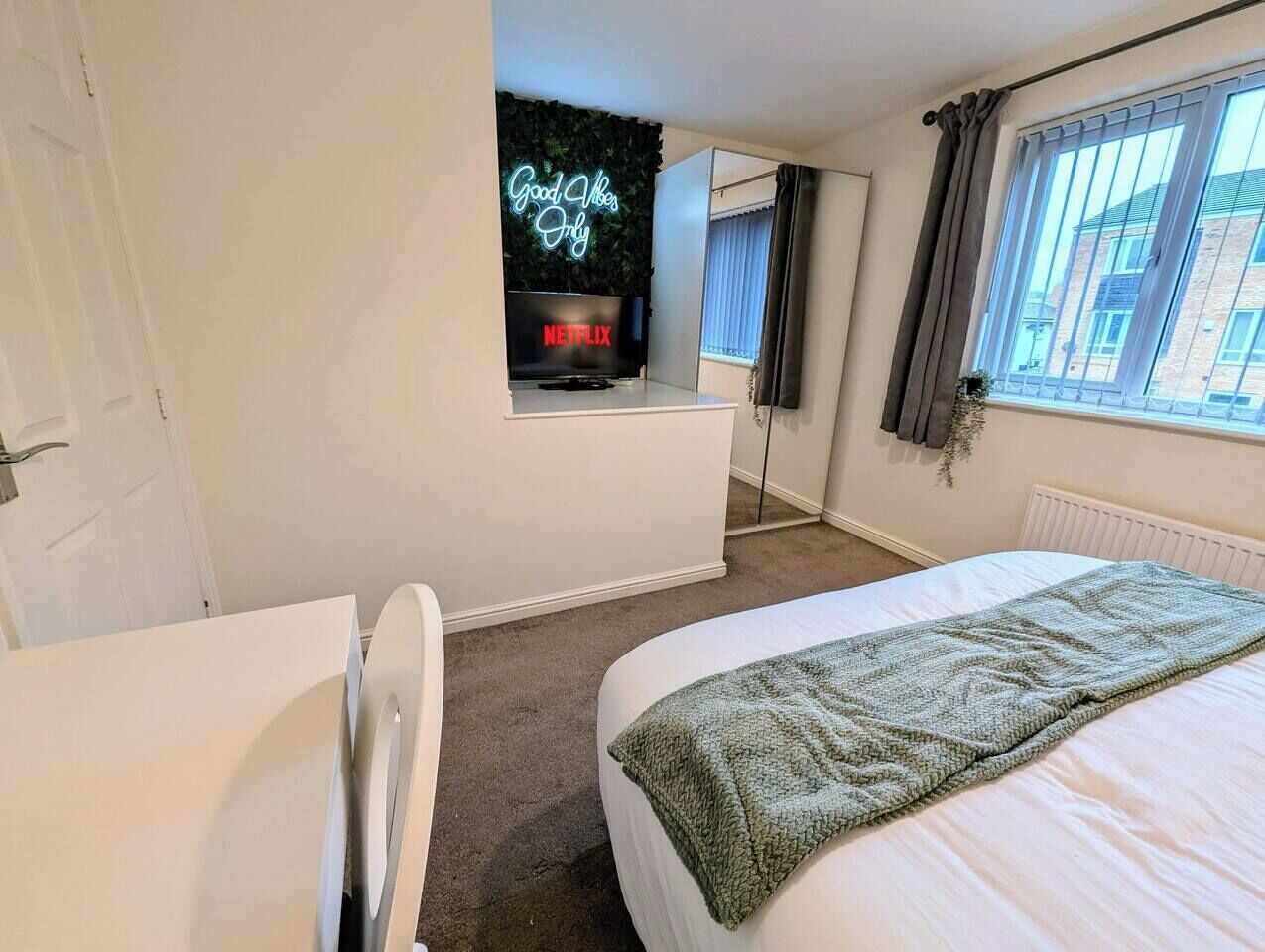 A bedroom with a bed, a desk, a neon sign, a window with curtains, and a radiator.