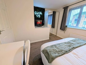 Ein Schlafzimmer mit Bett, Schreibtisch, einer Neonleuchtreklame, einem Fenster mit Vorhängen und einer Heizung.