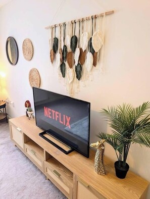 Ein Wohnzimmer mit Fernseher, der 'NETFLIX' anzeigt, einer Holz-Unterhaltungseinrichtung, einem wandmontierten Wandbehang aus Fransen und einem Topfpflanzen.