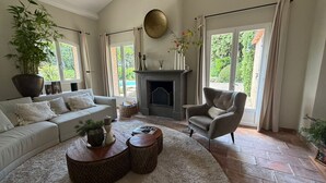 Ein Wohnzimmer mit einer Couch, Sesseln, Kamin, großen Fenstern und einer Topfpflanze.
