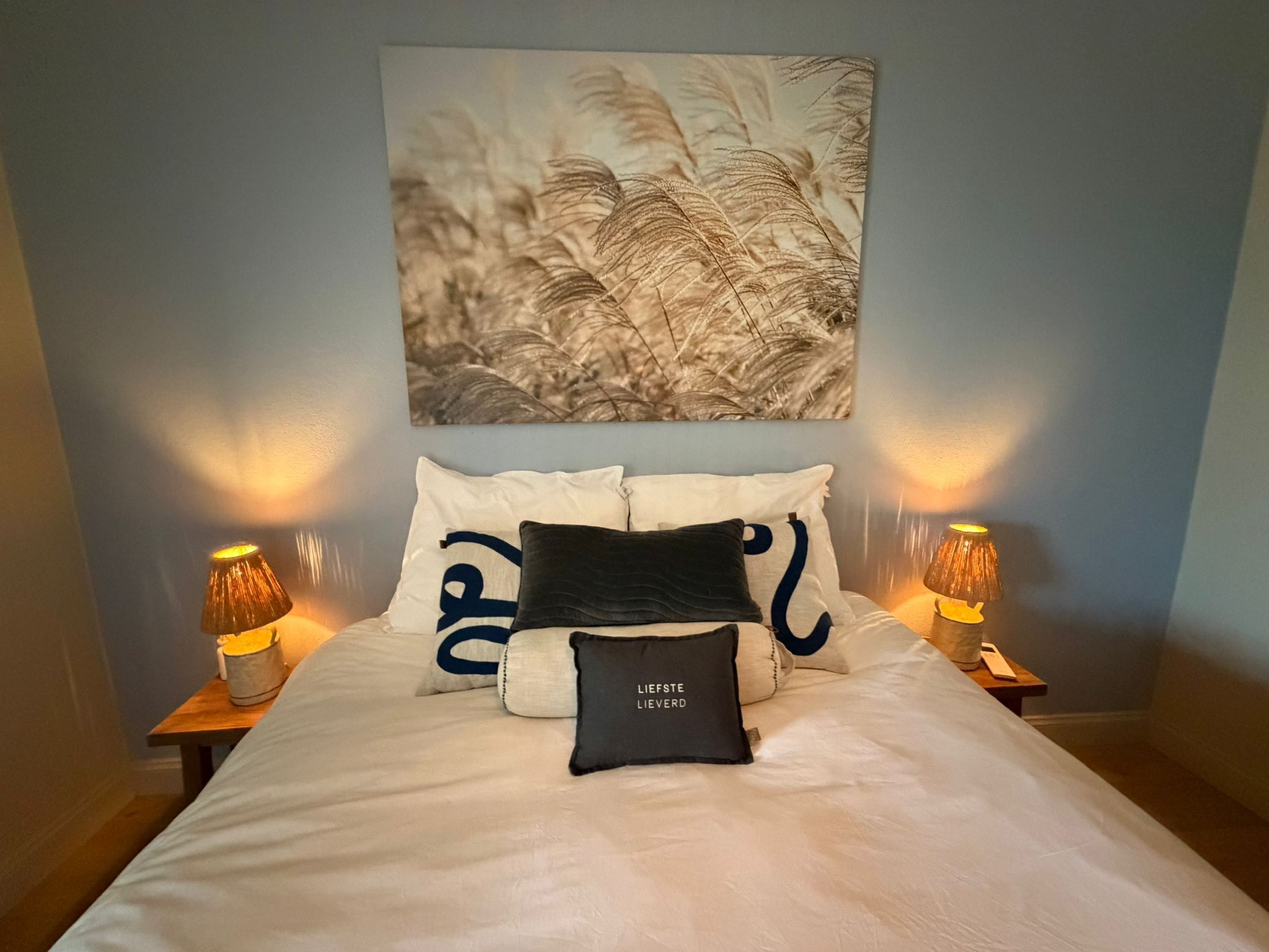 Een net opgemaakt bed met wit bedlinnen en twee decoratieve kussens, een kunstwerk aan de muur boven het bed, en twee bedlampen op houten nachtkastjes.