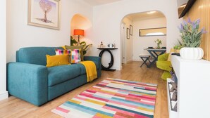 A living room with a teal sofa, colorful pillows, a multicolored rug, a framed artwork, a lamp, and a small table with a radio.