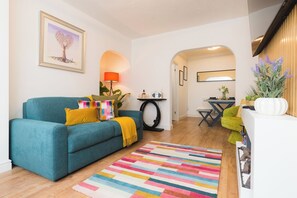 A living room with a teal sofa, colorful pillows, a multicolored rug, a framed artwork, a lamp, and a small table with a radio. - Heath Lodge - Stockingford, Nuneaton (Warwickshire)