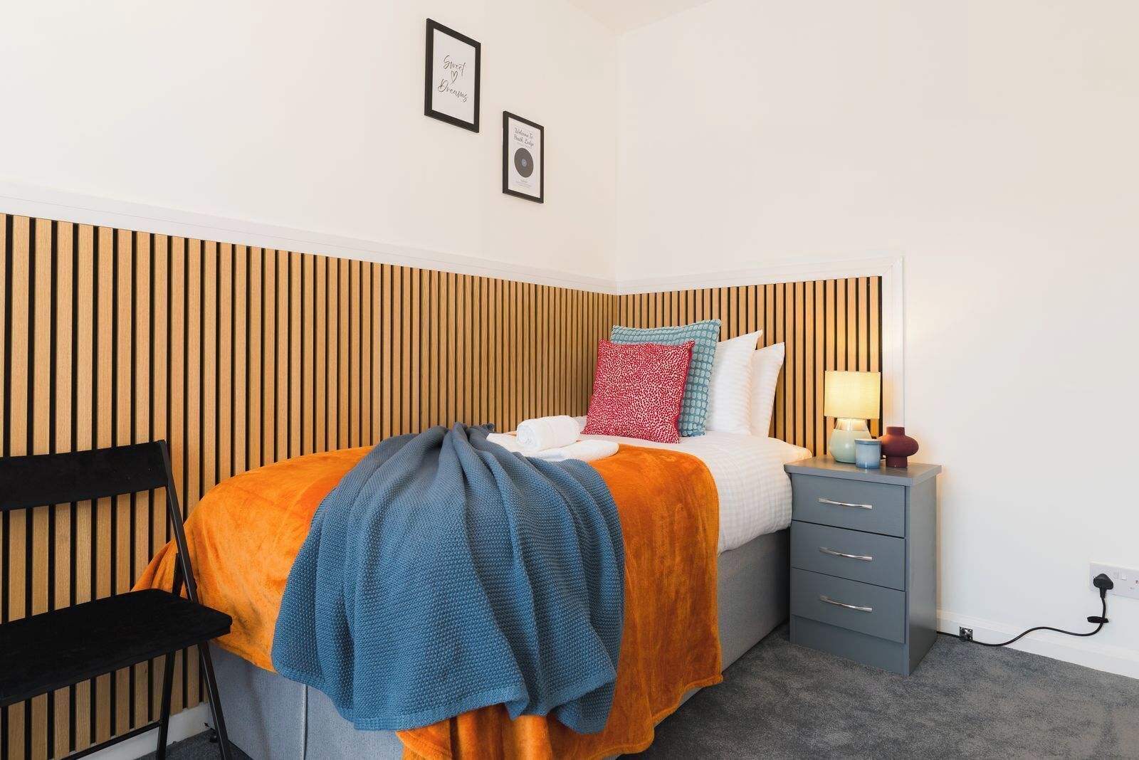 Un dormitorio ordenado con cama, mesita de noche, lámpara y una silla.