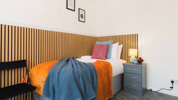 Un dormitorio ordenado con cama, mesita de noche, lámpara y una silla.