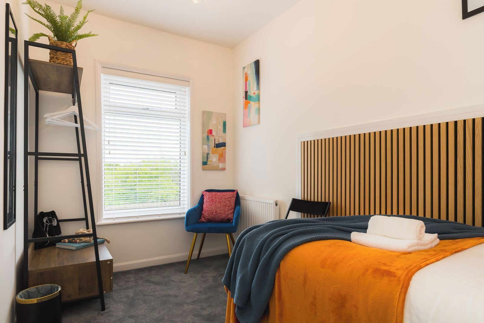 Ein Schlafzimmer mit einem Bett, einem Sessel, einer Leiter, einer Heizung, einem Fenster mit Jalousien und einem Wandbild.