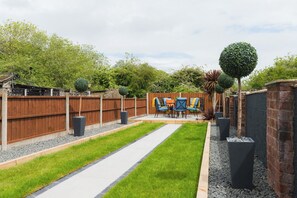 A backyard with a wooden fence, a paved pathway, a seating area with chairs and a table, and a well-maintained lawn. - Heath Lodge - Ideal for Families, Contractors & Professionals. (Warwickshire)