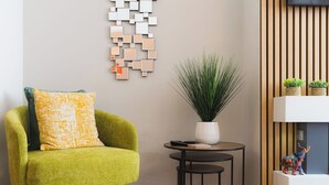 A green armchair with a patterned cushion, a small round table with a plant, and a wall-mounted abstract art piece.