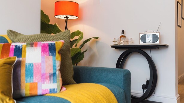 A living room with a sofa, colorful pillows, a lamp, a small table with a radio, and a plant. - Heath Lodge - Ideal for Families, Contractors & Professionals. (Warwickshire)