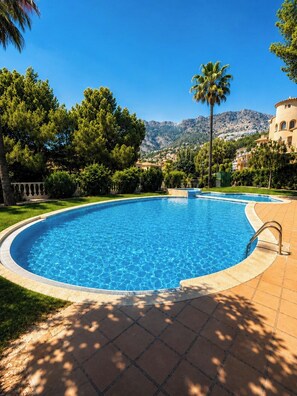 Ein klares, blaues Schwimmbecken mit einer gefliesten Terrasse, umgeben von viel Grün und Palmen, mit einer Bergkulisse im Hintergrund.