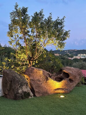 A landscaped garden with a tree, large rocks, and a grassy area. - Charming apartment in Porto Cervo, Sardegna. (Porto Cervo)