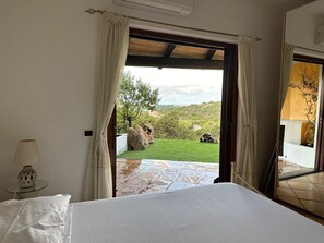 Quarto com cama, mesa com abajur e vista para um jardim através de uma porta aberta.