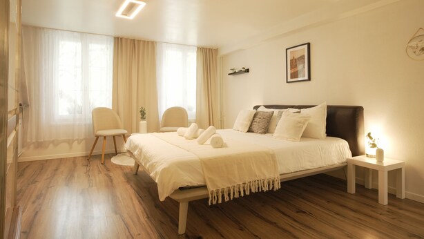 Un dormitorio con cama, mesitas de noche, una silla y una ventana con cortinas.