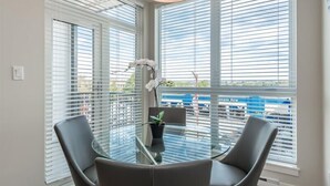 Una habitación con una mesa de vidrio rodeada de sillas, ventanas amplias con persianas y una planta en maceta.
