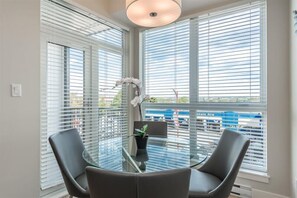 Ein Zimmer mit einem Glastisch, umgeben von Stühlen, großen Fenstern mit Jalousien und einer Zimmerpflanze.