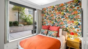 A bedroom with a large window, a bed with an orange bedspread, a colorful floral wallpaper, and a nightstand with a lamp.
