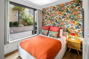 Ein Schlafzimmer mit einem großen Fenster, einem Bett mit orangefarbener Bettdecke, farbenfrohem Blumenwallpaper und einem Nachttisch mit Lampe.