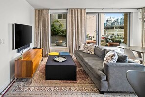 Ein modernes Wohnzimmer mit Flachbildfernseher, einer Couch, einem Couchtisch und Blick auf die Stadt durch große Fenster.