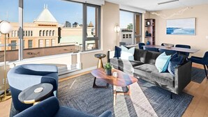 Ein modernes Wohnzimmer mit Sofa, Couchtisch und Essbereich, große Fenster mit Blick auf ein Gebäude mit der Aufschrift 'HUDSON'S BAY'.