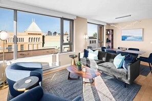 A modern living room with a sofa, coffee table, and dining area, large windows with a view of a building labeled 'HUDSON'S BAY'. - New! Ocean and Mountain View King Suite (Victoria)
