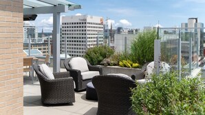 Eine Dachterrasse mit Korbmöbeln, einer Glasumlaufung und Blick auf die Stadt.