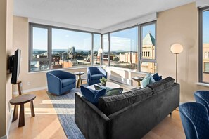 A modern living room with a large window, a black sofa, blue armchairs, and a small coffee table. - New! Ocean and Mountain View King Suite (Victoria)