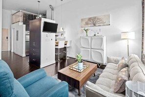 Ein modernes Wohnzimmer mit einer Couch, einem Couchtisch und einem Fernseher. Es gibt eine Küchenzeile mit Schränken und einem Esstisch.